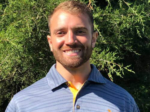 man with beard wearing blue striped polo shirt in front of a bush